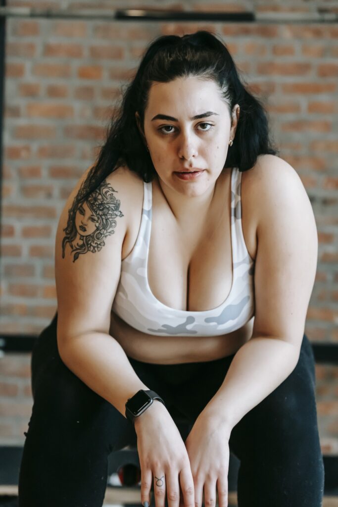 Serious young plus size tattooed female athlete with long dark hair in activewear recreating on gym bench after intense workout